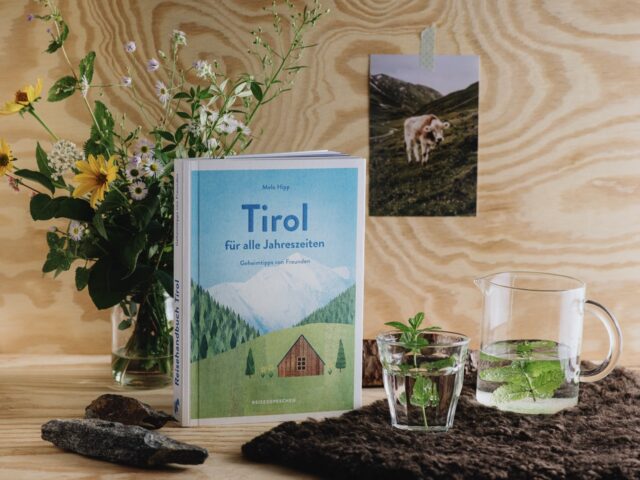Tirol Reiseführer auf einem Holztisch, daneben ein Blumenstrauß, Wasserkaraffe mit Minze und ein Glas; im Hintergrund ein Foto einer Kuh an der Holzwand – stimmungsvolle Szene mit alpinem Flair.