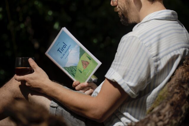 Mann in sommerlicher Kleidung liest den Tirol Reiseführer in dunkler Umgebung, Getränk in der Hand – ruhige Reiseszene mit Fokus auf das Buch “Tirol für alle Jahreszeiten”
