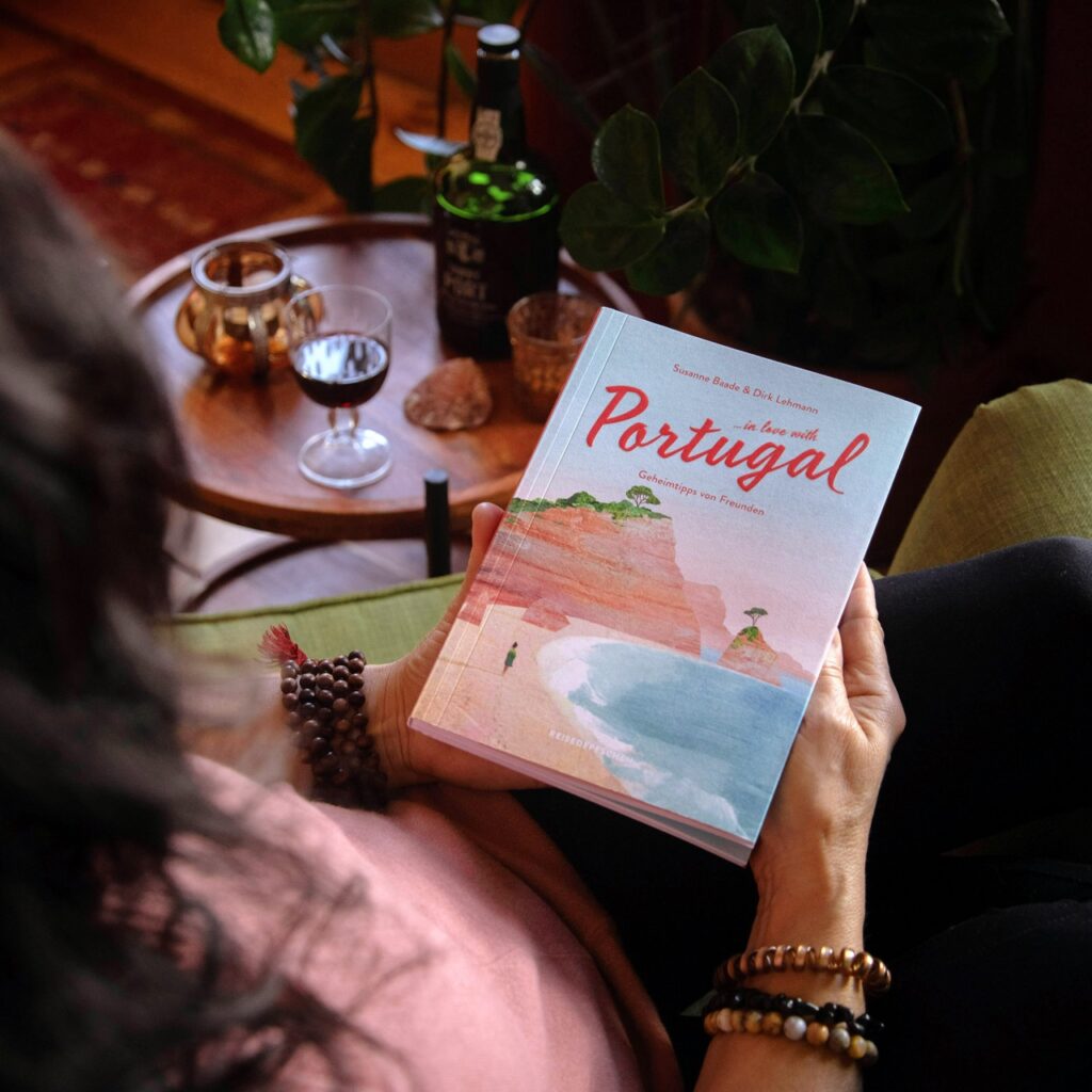 Portugal Reiseführer „..in love with Portugal“ in der Hand gehalten, im Hintergrund ein Tisch mit Wein – stimmungsvolle Szene zur Einstimmung auf eine Portugal-Reise.
