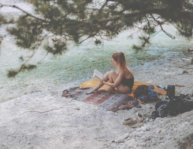 Eine Frau liest ein Buch und sitzt auf weißem, steinigem Ufer am Fluss Mala Pišnica, von hinten fotografiert, mit Blick auf grüne Bäume – ruhige Naturszene im Triglav-Nationalpark